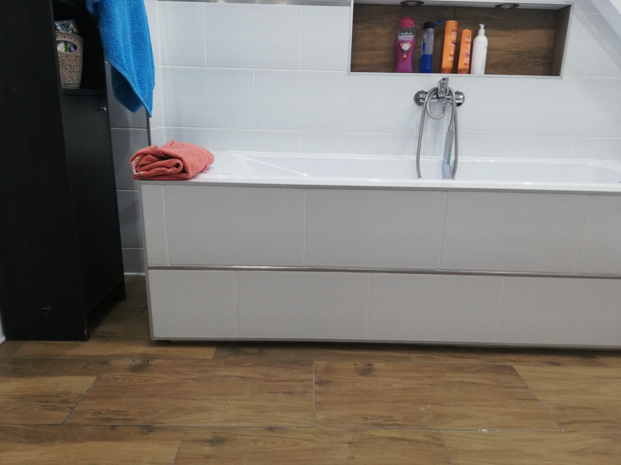 Nowoczesna łazienka z białą wanną obudowaną płytkami, drewnianą podłogą i wnęką z kosmetykami, uzupełniona o kolorowe ręczniki dla ożywienia wnętrza.