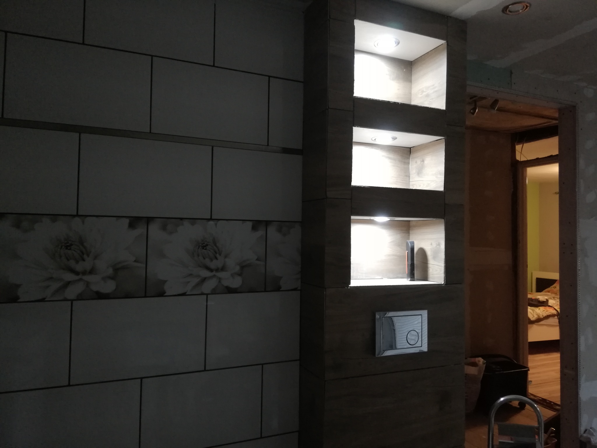 Ściana łazienki z płytkami w kwiatowy wzór i kolumna z wnękami podświetlonymi LED, widoczny przycisk spłuczki i fragment pokoju w tle, etap prac wykończeniowych.