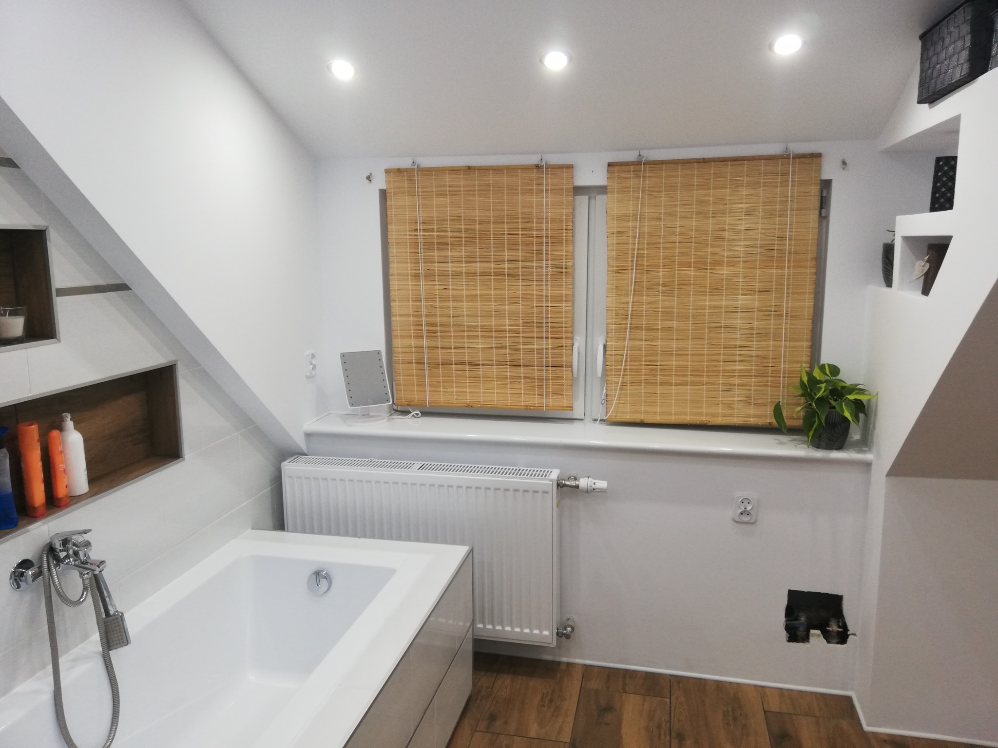 Biała wanna w łazience na poddaszu z drewnianą podłogą, bambusowymi roletami w oknach i odsłoniętą instalacją elektryczną.