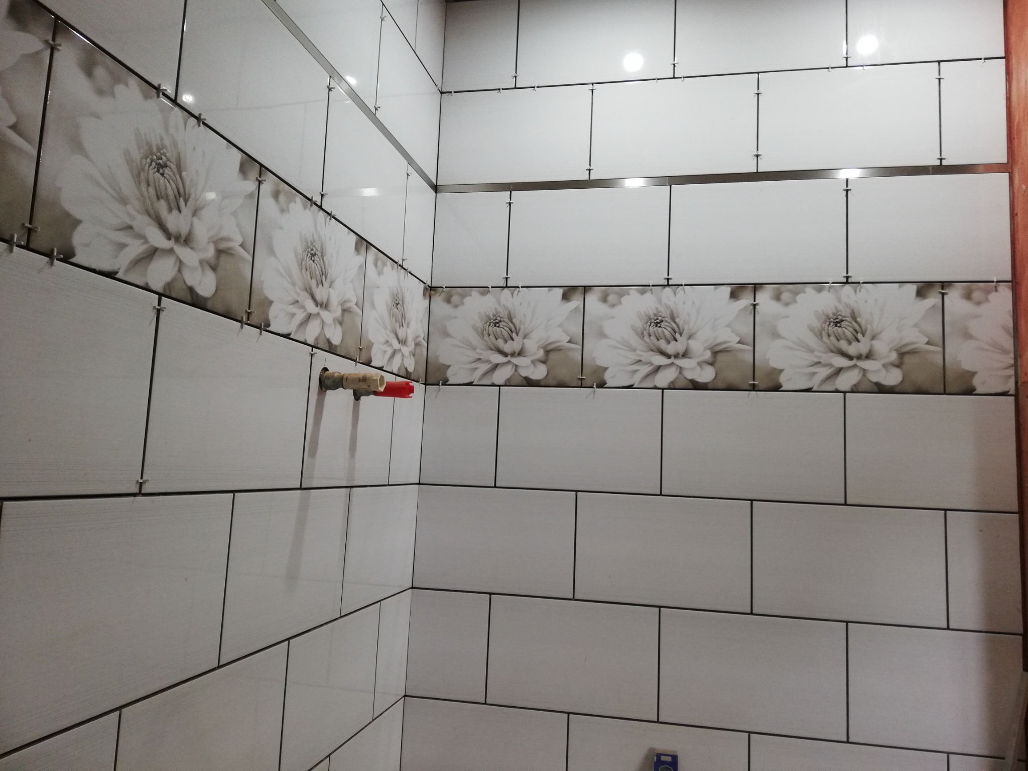 Ściany w trakcie układania płytek ceramicznych z dekoracyjnym pasem z motywem kwiatowym, widoczne krzyżyki dystansowe i rura z zaworem.