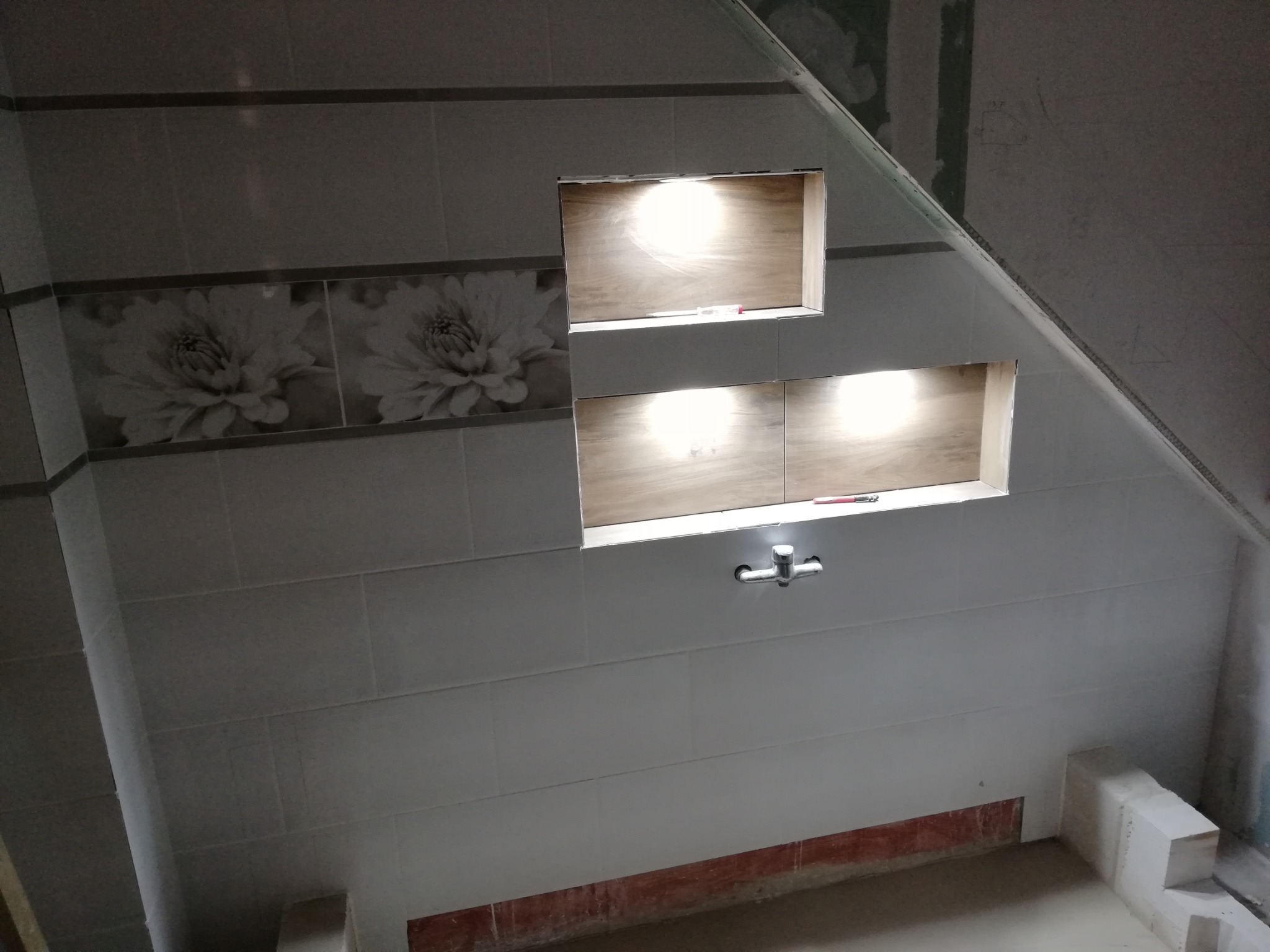 Ściana łazienki w trakcie wykańczania, widoczne białe płytki z dekoracyjnym pasem kwiatowym, wnęki oświetleniowe i surowy stan podłogi.