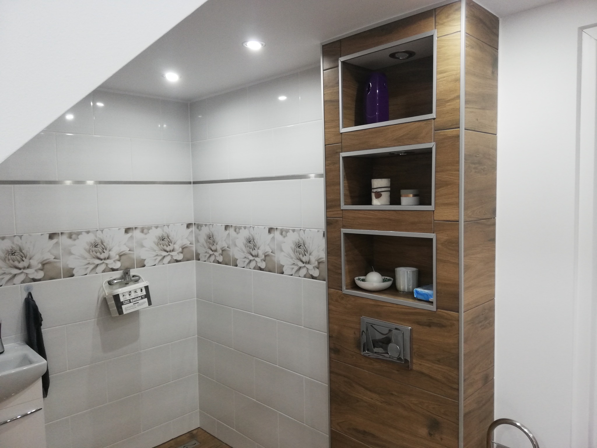 Wykończona łazienka w Gwieździnie: białe płytki z motywem kwiatowym, zabudowa z drewnopodobnych paneli z wnękami na dekoracje i akcesoria łazienkowe, podwieszana miska WC.