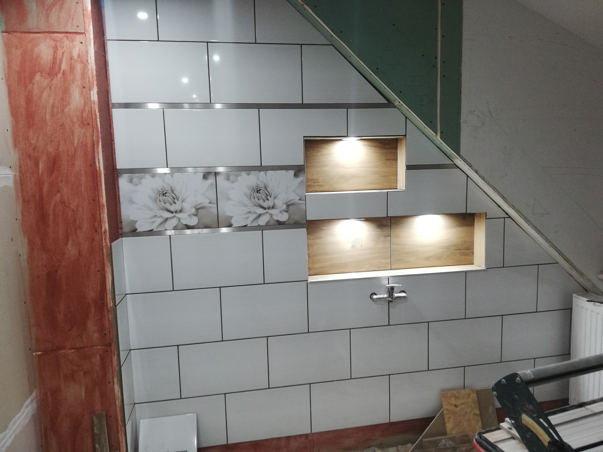 Wykończenie łazienki na poddaszu: białe płytki z motywem kwiatowym, wnęki z oświetleniem, widoczna konstrukcja z płyty kartonowo-gipsowej i narzędzie do cięcia płytek.