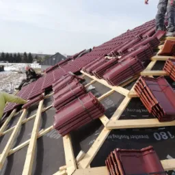 Montaż bordowej dachówki ceramicznej na drewnianej konstrukcji dachu, widoczni dekarze w trakcie pracy, czarna membrana dachowa z widocznym nadrukiem, w tle krajobraz z budynkami.