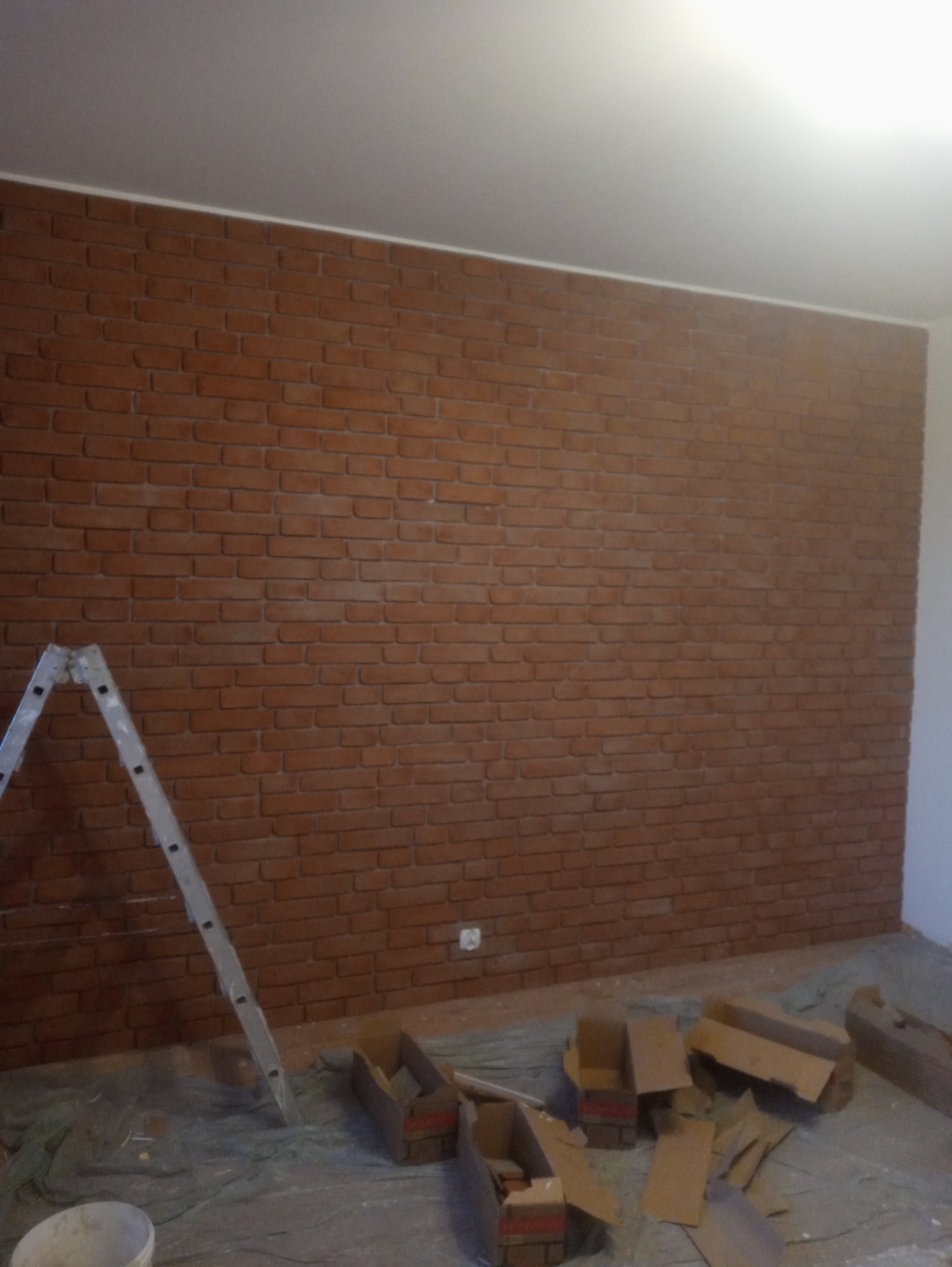 Ściana wyłożona cegłą dekoracyjną w odcieniach brązu, widoczna drabina malarska i kartony po płytkach na podłodze zabezpieczonej folią.