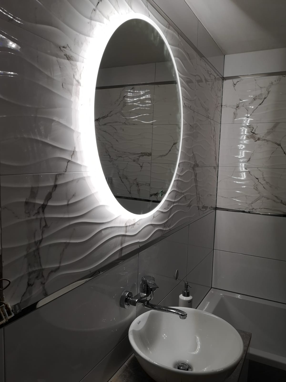 Nowoczesna łazienka z umywalką nablatową, chromowaną baterią i podświetlanym lustrem owalnym na ścianie z płytkami imitującymi marmur z falistą strukturą.
