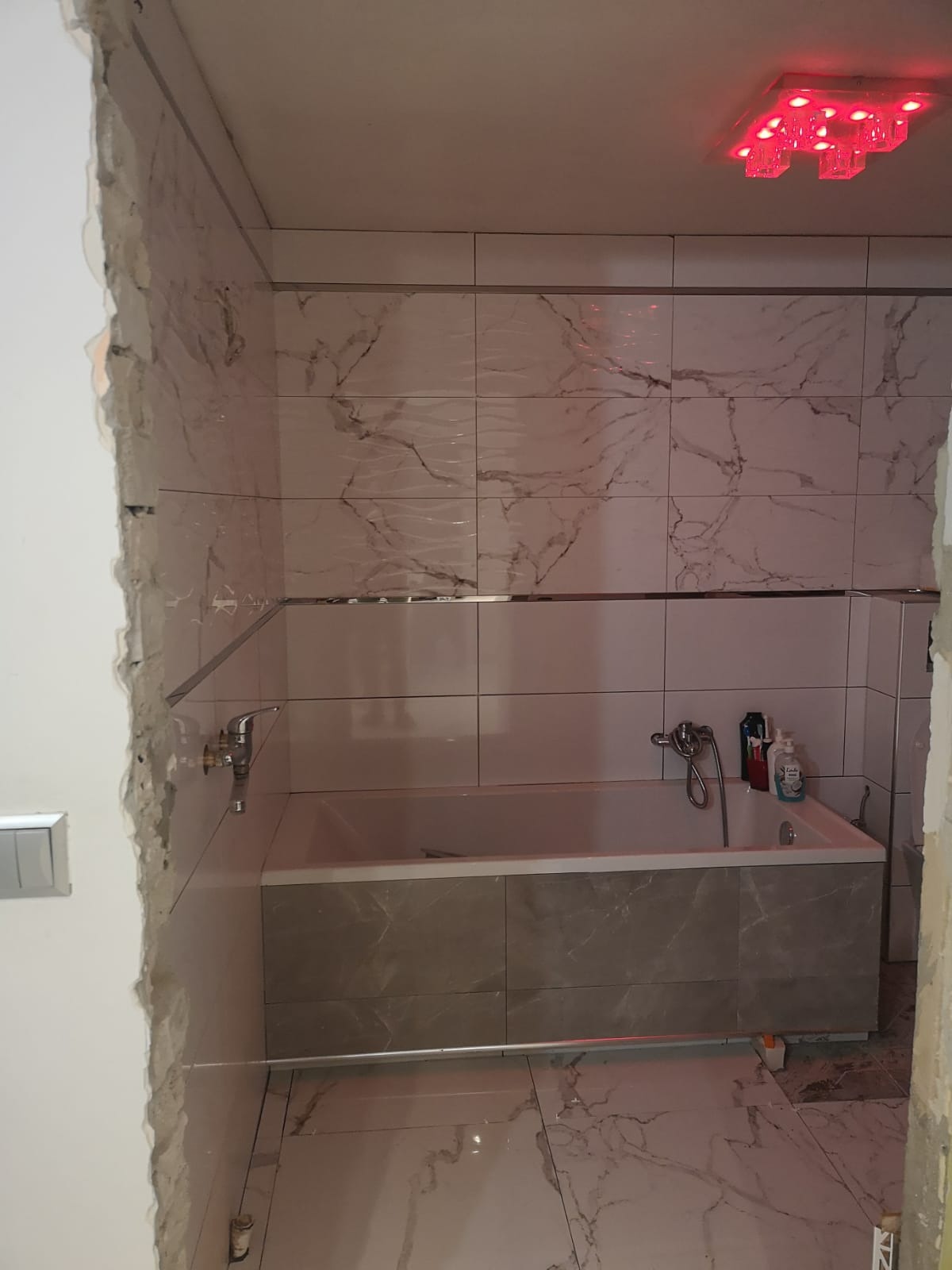 Remontowana łazienka z wanną obudowaną płytkami imitującymi marmur, ściany wyłożone płytkami z podobnym wzorem, widoczne niedokończone obrzeża i czerwone oświetlenie sufitowe.