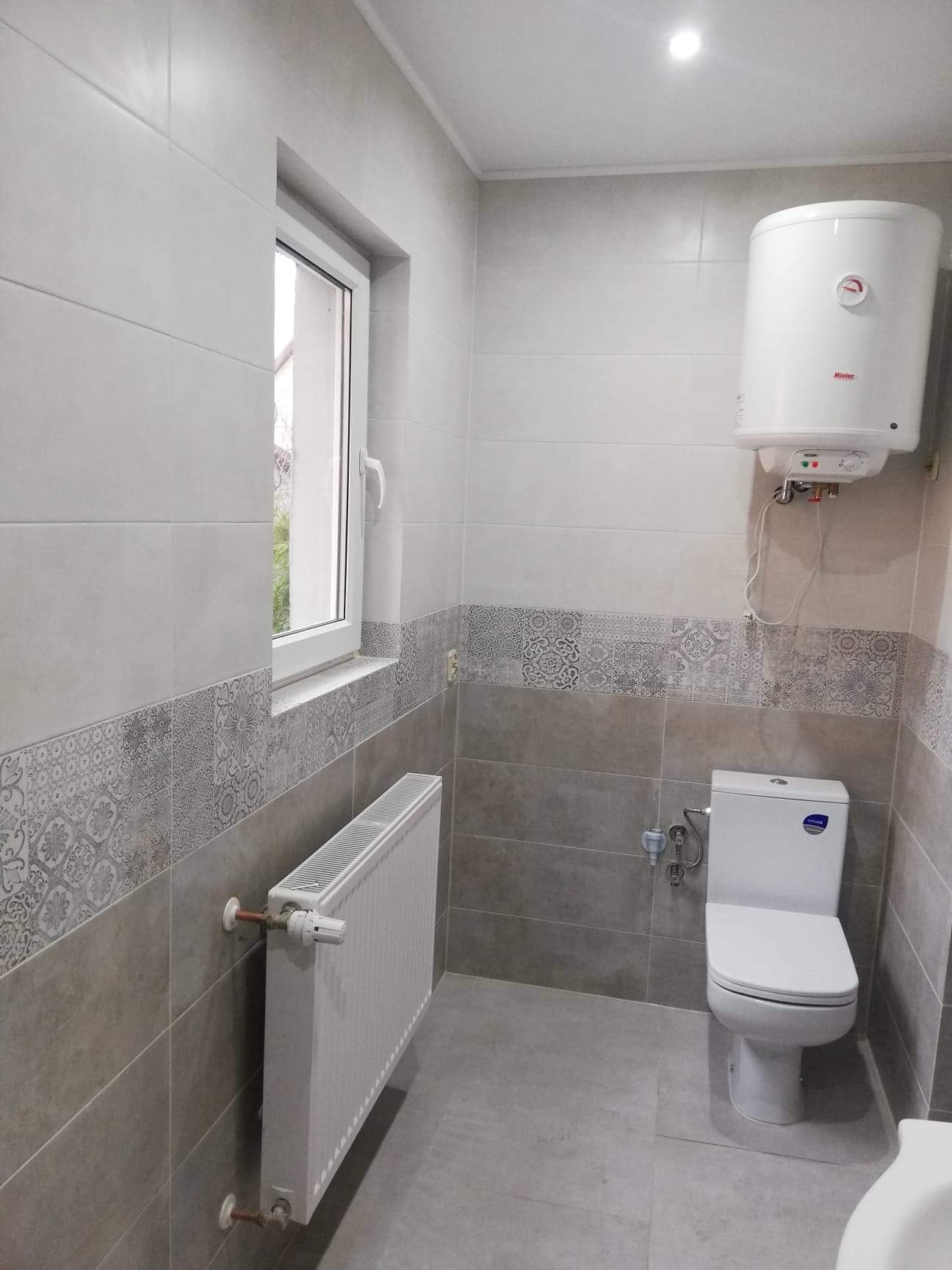 Wykończona łazienka w szaro-białej kolorystyce z białym bojlerem, grzejnikiem i toaletą typu kompakt, z dekoracyjnym pasem płytek na ścianach.