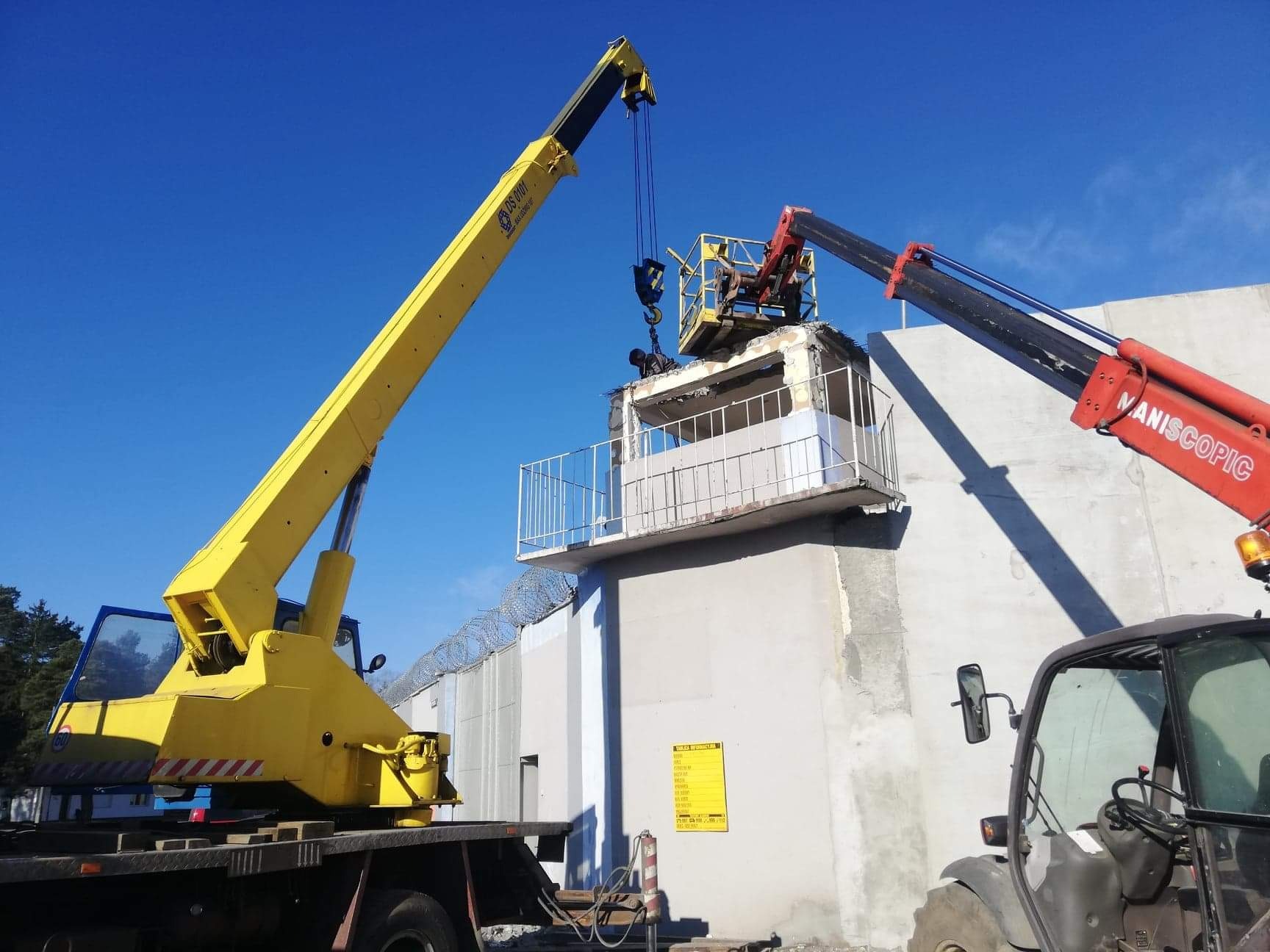 Demontaż betonowej konstrukcji z użyciem dwóch dźwigów na tle błękitnego nieba, widoczny fragment muru z drutem kolczastym.