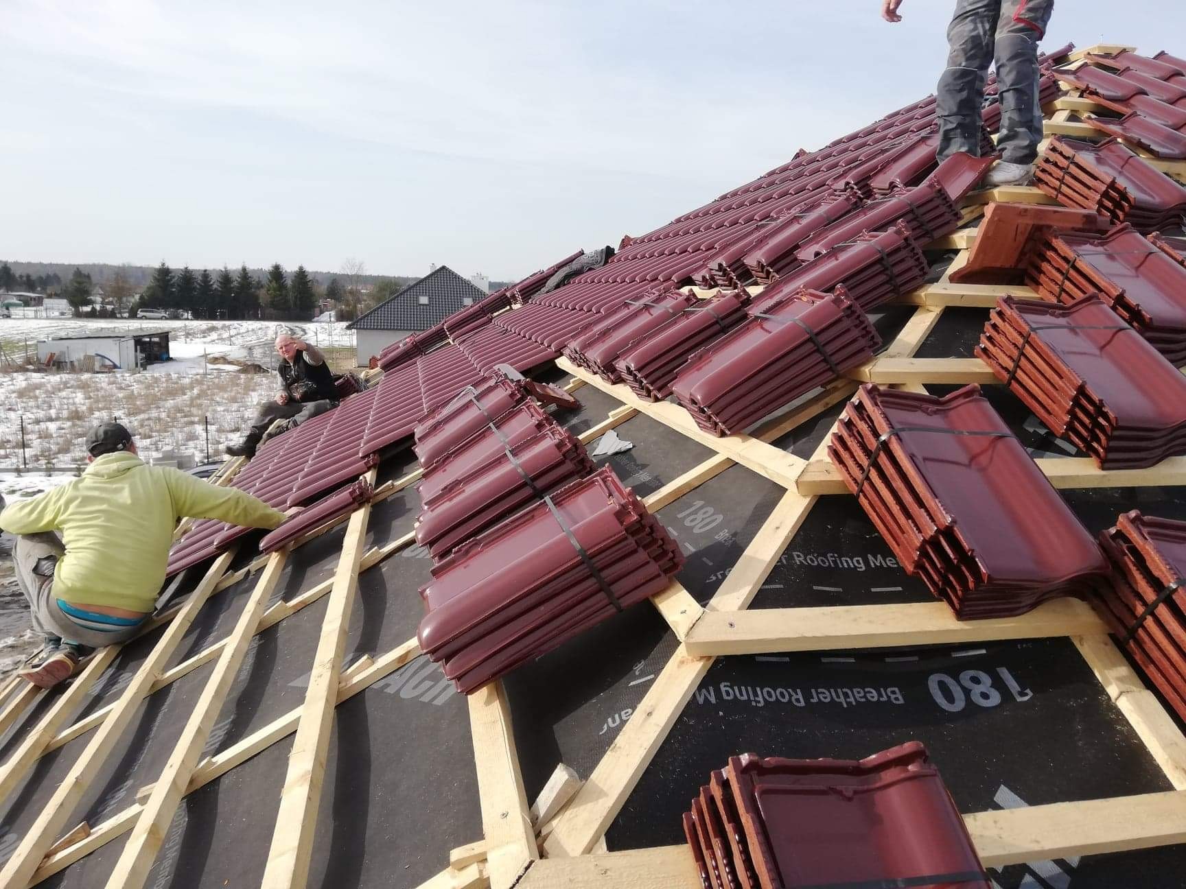 Montaż bordowej dachówki ceramicznej na drewnianej konstrukcji dachu, widoczni dekarze w trakcie pracy, czarna membrana dachowa z widocznym nadrukiem, w tle krajobraz z budynkami.