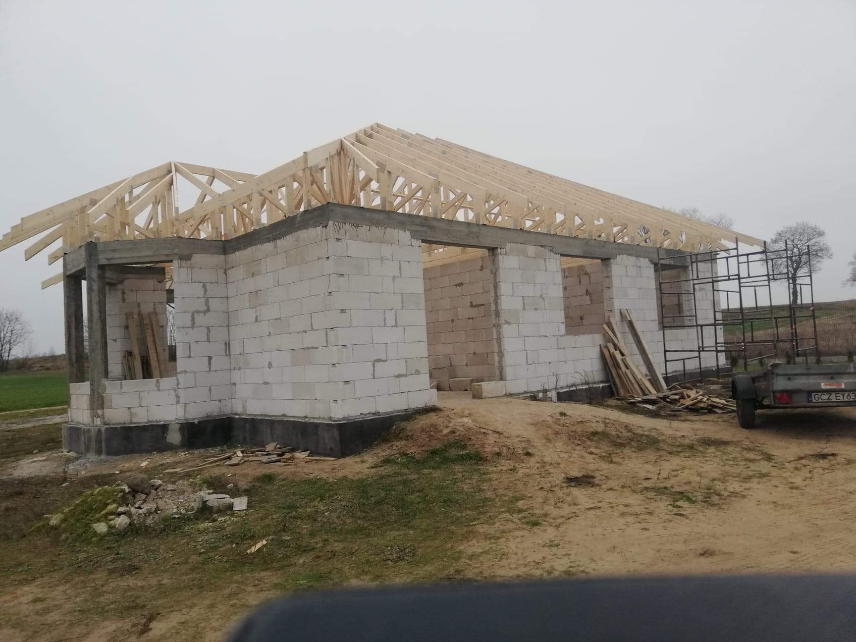 Surowy stan budowy domu jednorodzinnego z bloczków, widoczna więźba dachowa, teren budowy z przyczepką i stertą desek, pochmurne niebo.