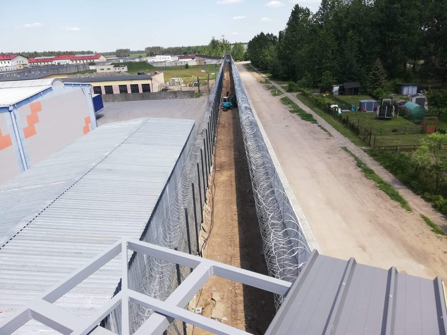 Widok z góry na długi korytarz z drutem kolczastym po obu stronach, mała koparka w oddali, obok droga gruntowa i zabudowania.
