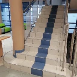 Klatka schodowa w biurowcu z kolumną obudowaną płytkami i metalowymi barierkami, wyłożona jasnymi płytkami z niebieskimi pasami na stopniach.