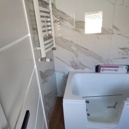 Yaroslav Zakhidnyy - Nowoczesna łazienka z białą wanną z przeszklonymi drzwiczkami, grzejnikiem drabinkowym i marmurkowymi płytkami ściennymi, widok na brązową podłogę imitującą drewno oraz fragment czarnej klamki...