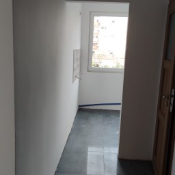 Yaroslav Zakhidnyy - Wnętrze łazienki w trakcie remontu: szare płytki podłogowe, białe ściany z wyprowadzonymi instalacjami wodnymi, widok na okno z fragmentem zabudowy mieszkalnej na zewnątrz, częściowo widoczne...