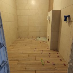 Yaroslav Zakhidnyy - Wnętrze łazienki w trakcie prac wykończeniowych; na podłodze ułożone płytki imitujące drewno, z widocznymi krzyżykami dystansowymi i elementami poziomowania; na ścianach beżowe płytki kwadratowe...