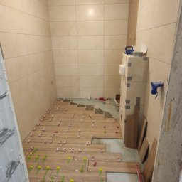 Yaroslav Zakhidnyy - Układanie płytek imitujących drewno na podłodze w łazience, widoczne krzyżyki dystansowe w kolorach czerwonym i zielonym, ściany wyłożone jasnymi płytkami, surowy stan wykończenia.