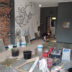 Yaroslav Zakhidnyy - Układanie płytek podłogowych w lokalu w Osnabrück, widoczny glazurnik przy pracy, zaplecze z umywalką i toaletą w tle, ściana z motywem ośmiornicy, narzędzia i materiały budowlane porozkładane...