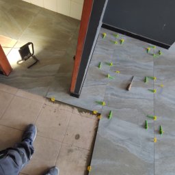 Yaroslav Zakhidnyy - Układanie szarej płytki podłogowej z użyciem systemu poziomowania płytek (żółte i zielone kliny) w łazience, widoczny fragment starej podłogi, narzędzie glazurnicze i oświetlenie robocze.