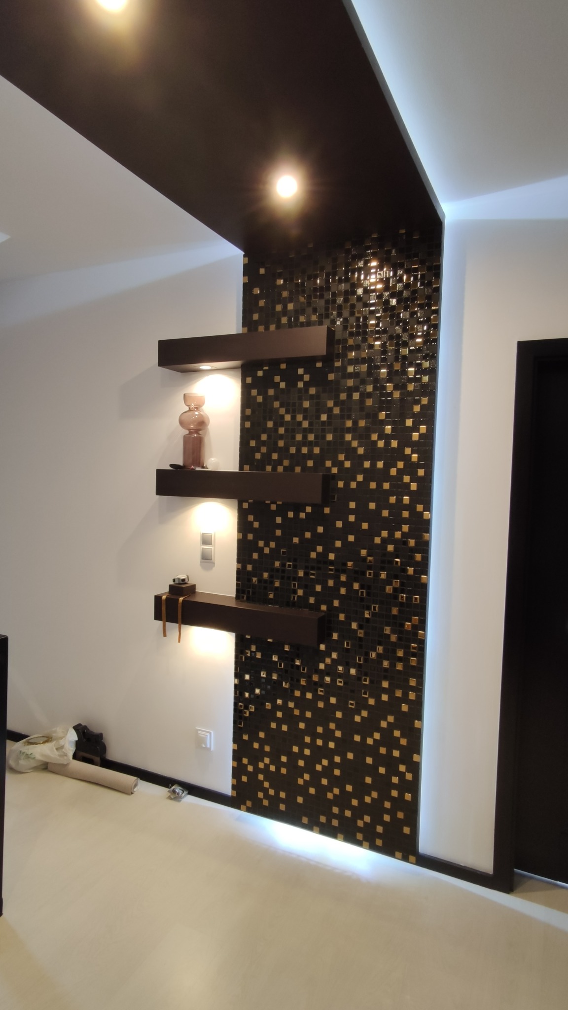 Ściana akcentowa z mozaiki w kolorze czarnym i złotym, podświetlane półki z dekoracjami, czarny pas sufitowy z wbudowanym oświetleniem, jasna podłoga.