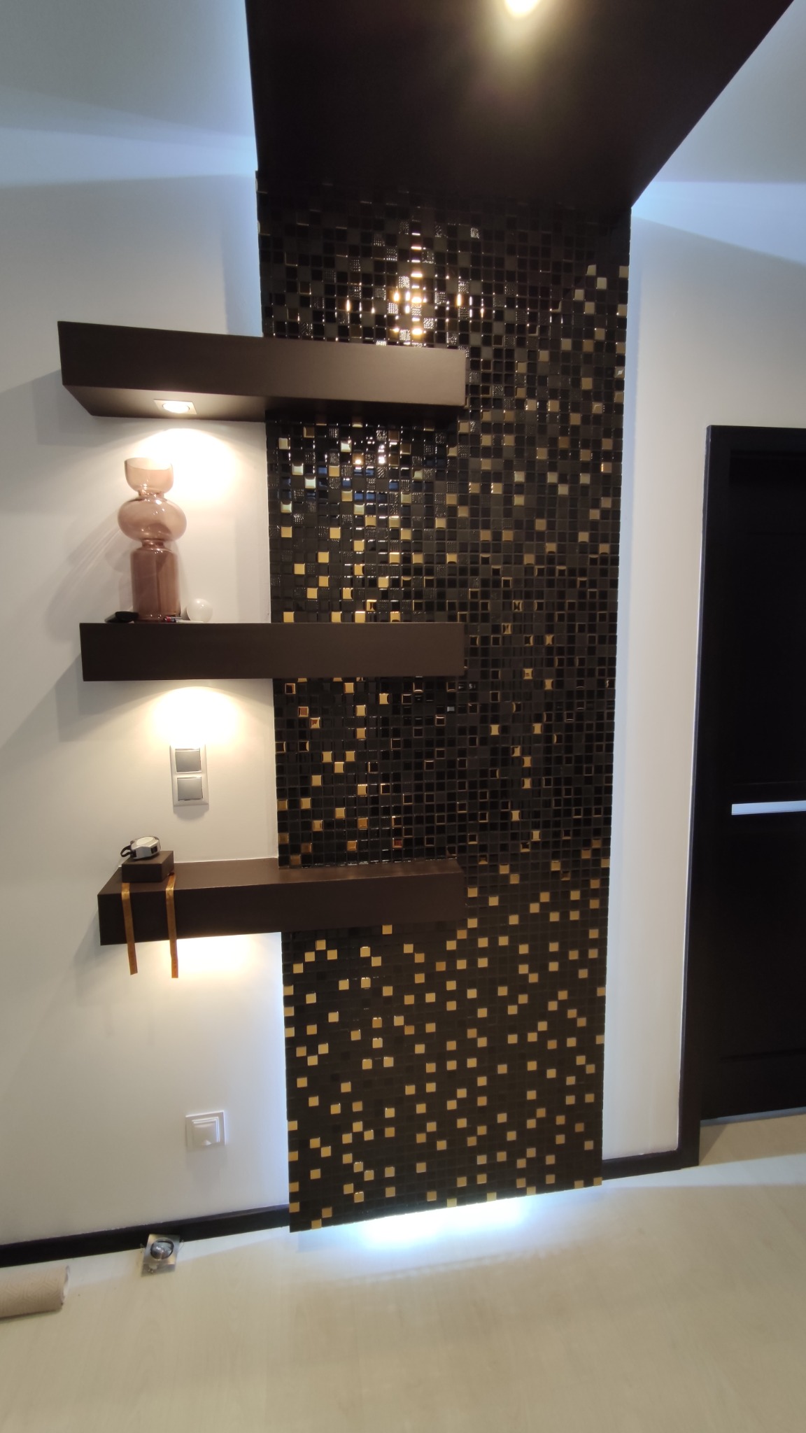 Ściana wykończona czarną mozaiką z złotymi akcentami, oświetlona od góry i dołu, z trzema półkami w kolorze ciemnego brązu, na których stoją dekoracje i wbudowane są lampki punktowe.