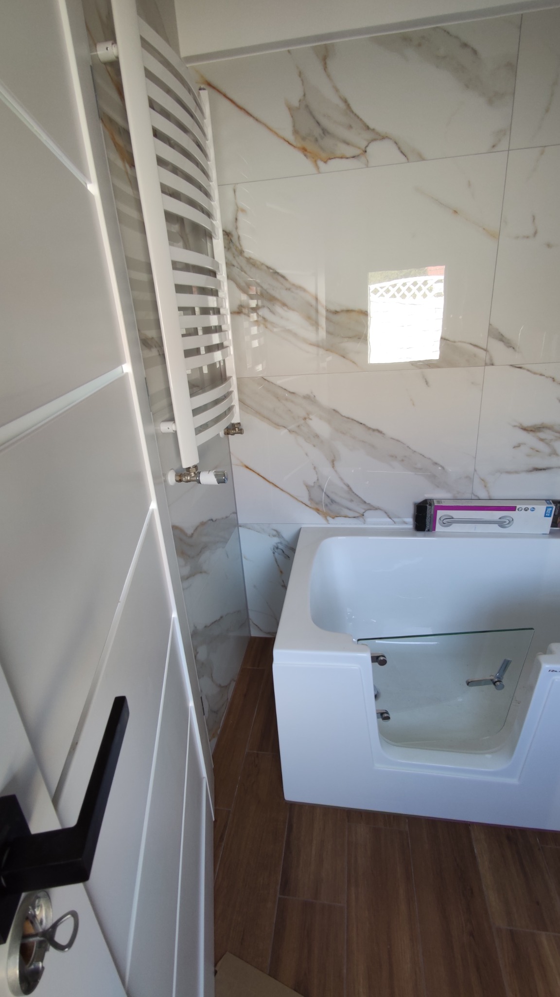 Nowoczesna łazienka z białą wanną z przeszklonymi drzwiczkami, grzejnikiem drabinkowym i marmurkowymi płytkami ściennymi, widok na brązową podłogę imitującą drewno oraz fragment czarnej klamki...