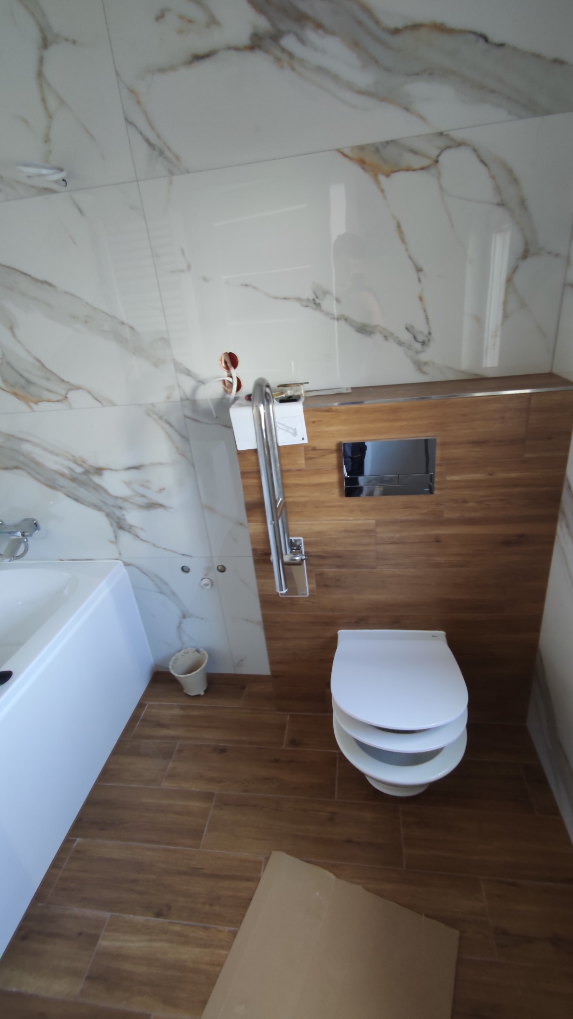 Wykończona łazienka z marmurowymi płytkami na ścianach, drewnopodobną podłogą, podwieszaną toaletą i chromowaną poręczą dla niepełnosprawnych.