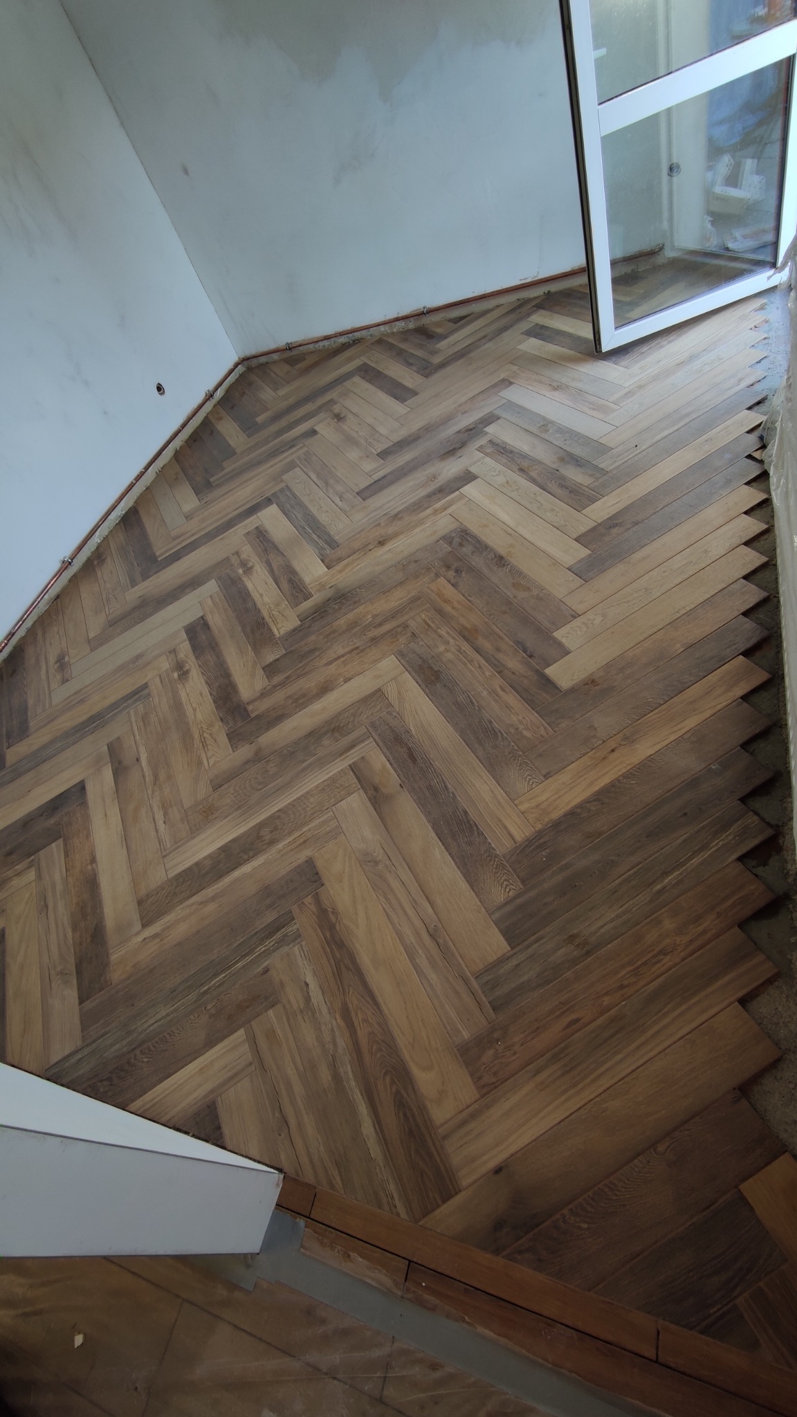 Układanie parkietu w jodełkę z widocznymi niedociągnięciami przy ścianie i ościeżnicy okiennej, surowy stan wykończenia.