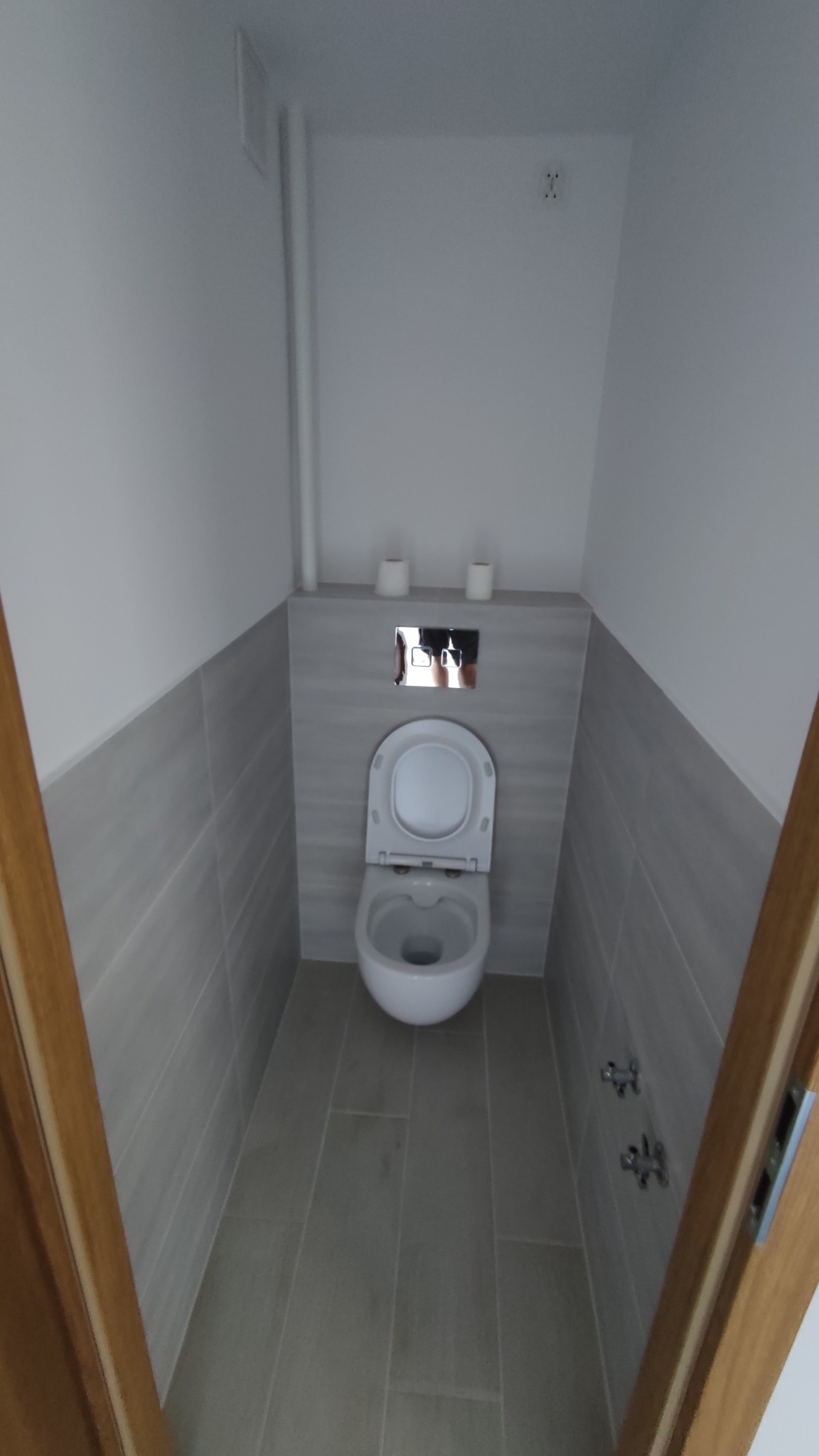 Wąska toaleta po remoncie, wykończona szarymi płytkami imitującymi drewno, z podwieszaną muszlą klozetową i minimalistycznym panelem sterującym spłukiwaniem.