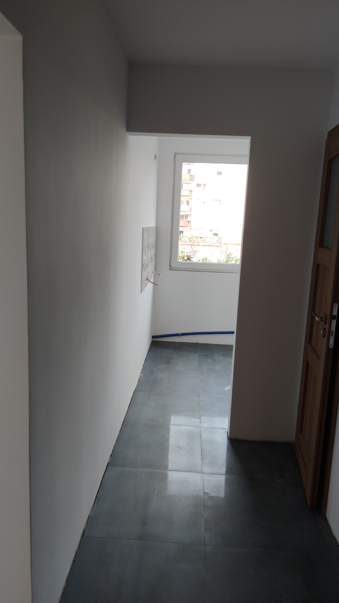 Wnętrze łazienki w trakcie remontu: szare płytki podłogowe, białe ściany z wyprowadzonymi instalacjami wodnymi, widok na okno z fragmentem zabudowy mieszkalnej na zewnątrz, częściowo widoczne...