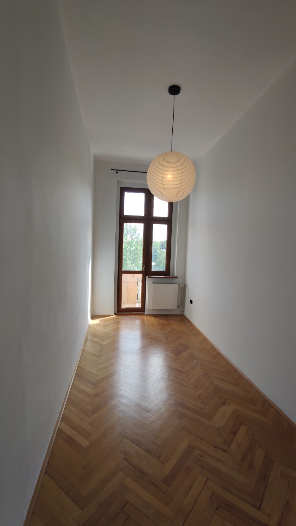Wnętrze wąskiego pokoju po remoncie z parkietem ułożonym w jodełkę, świeżo pomalowanymi na biało ścianami i papierową lampą wiszącą.