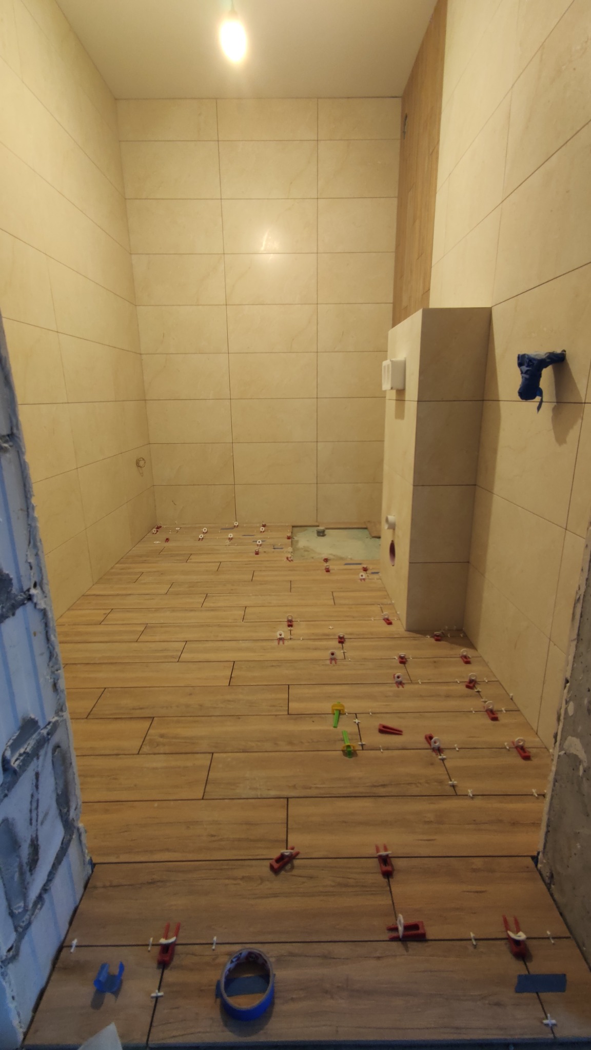 Wnętrze łazienki w trakcie prac wykończeniowych; na podłodze ułożone płytki imitujące drewno, z widocznymi krzyżykami dystansowymi i elementami poziomowania; na ścianach beżowe płytki kwadratowe...