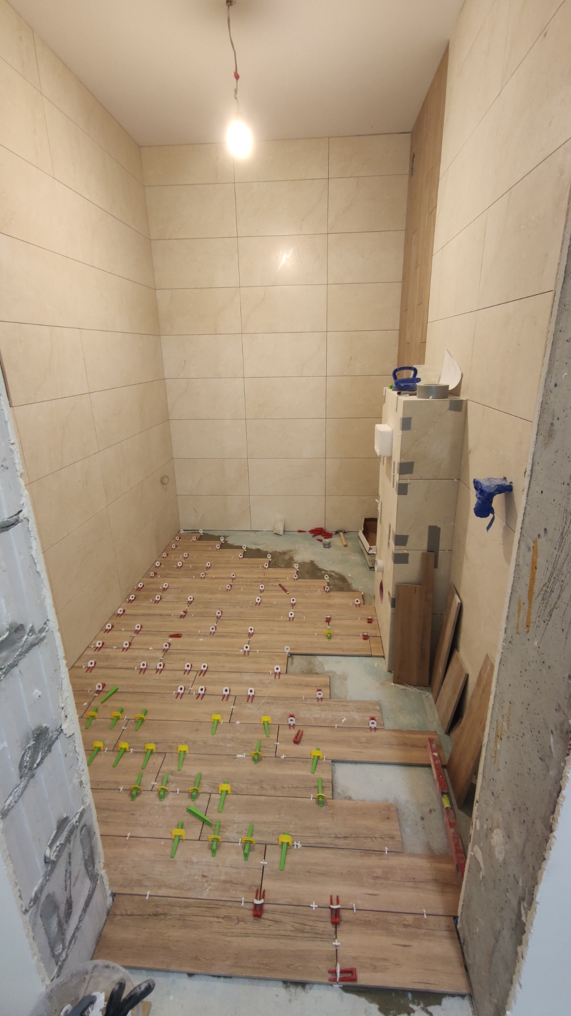 Układanie płytek imitujących drewno na podłodze w łazience, widoczne krzyżyki dystansowe w kolorach czerwonym i zielonym, ściany wyłożone jasnymi płytkami, surowy stan wykończenia.
