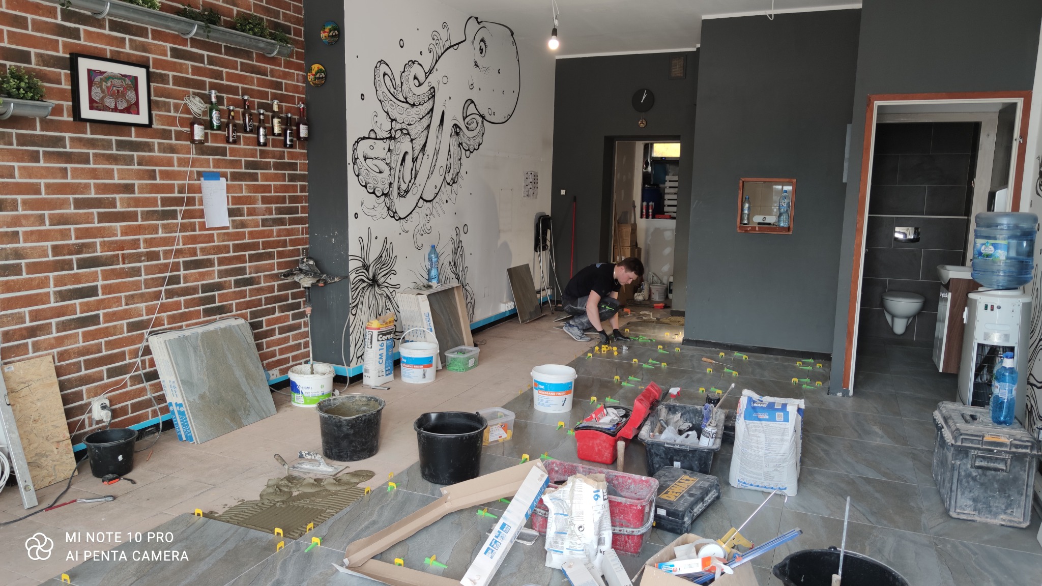 Układanie płytek podłogowych w lokalu w Osnabrück, widoczny glazurnik przy pracy, zaplecze z umywalką i toaletą w tle, ściana z motywem ośmiornicy, narzędzia i materiały budowlane porozkładane...