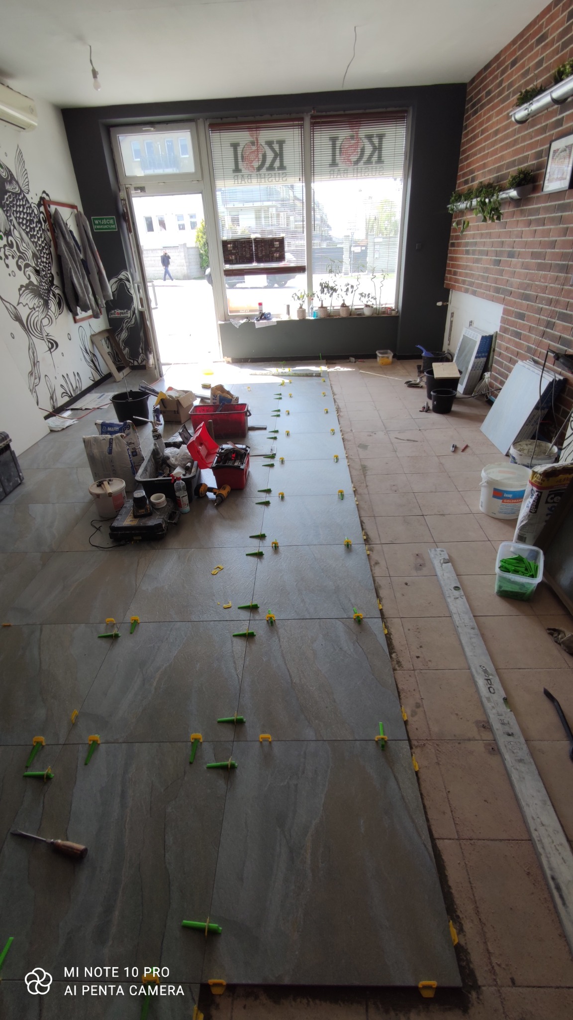 Wnętrze lokalu z częściowo ułożoną podłogą z płytek ceramicznych w odcieniach szarości, widoczne narzędzia, poziomica i plastikowe krzyżyki dystansowe między płytkami. Po prawej stronie widoczny...