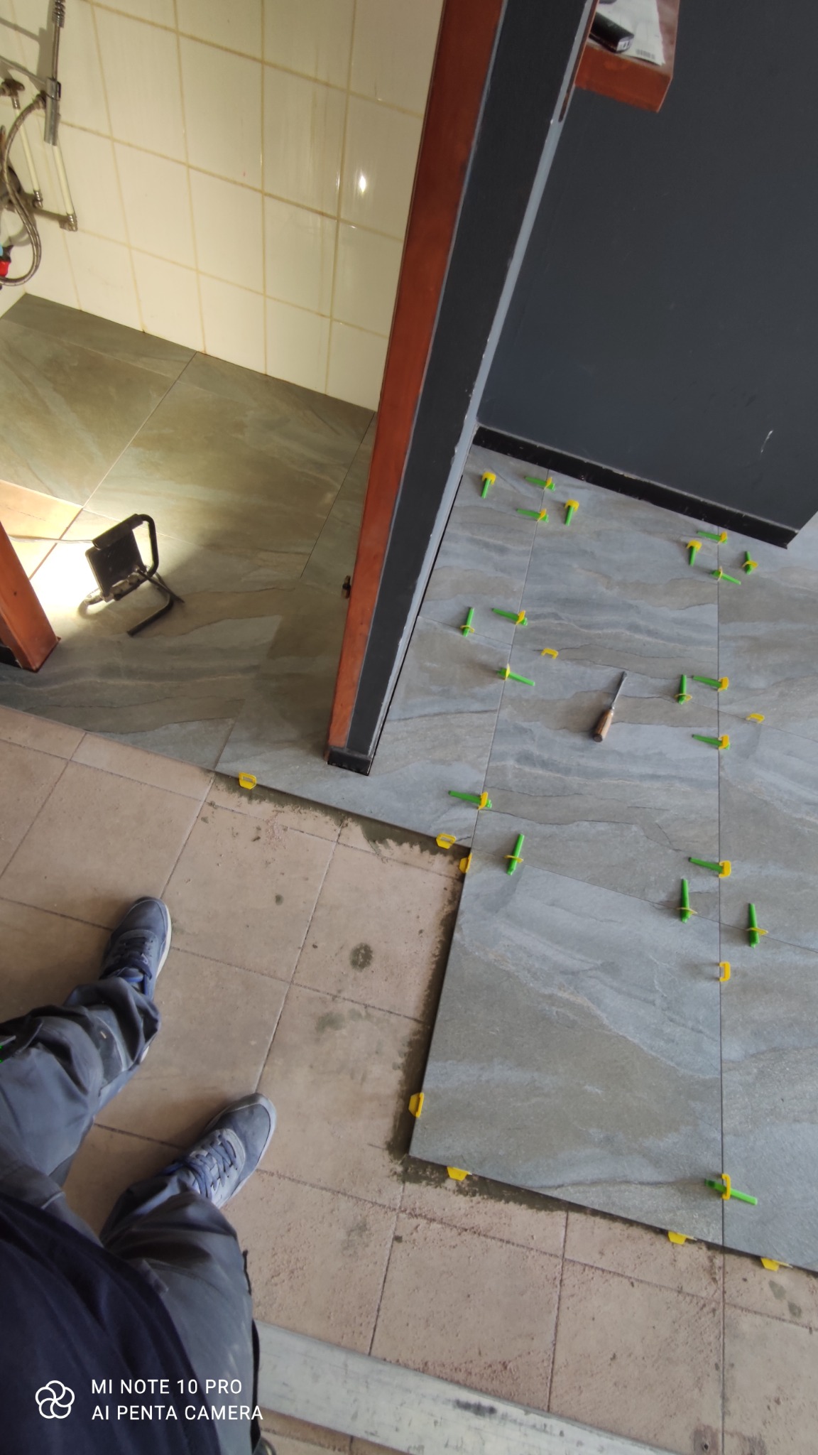 Układanie szarej płytki podłogowej z użyciem systemu poziomowania płytek (żółte i zielone kliny) w łazience, widoczny fragment starej podłogi, narzędzie glazurnicze i oświetlenie robocze.