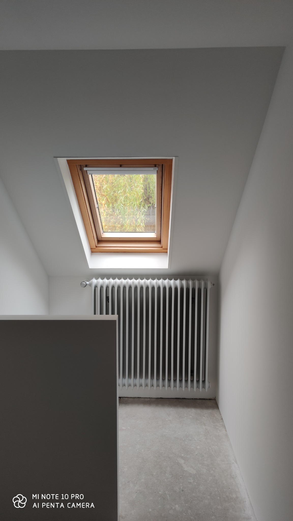 Poddasze po remoncie, widoczne białe ściany, okno dachowe z drewnianą ramą, grzejnik pomalowany na biało i surowa betonowa posadzka przed położeniem wykończenia.