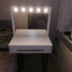 Biała toaletka z lustrem i oświetleniem LED, ustawiona w pokoju obok okna z firanką, widoczny fragment łóżka i grzejnika