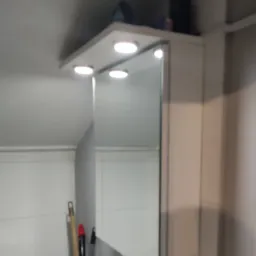 Biała szafka łazienkowa z lustrem i oświetleniem LED, zamontowana na ścianie nad umywalką, widok z boku, z elementami wyposażenia łazienki w tle.