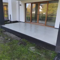 Nowoczesny taras z jasnoszarej płyty, obrzeża z ciemnej kostki, widok na ogród przez duże przesuwne okna.