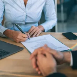 Osoba w błękitnej koszuli podpisuje dokument na drewnianym biurku, obok leży laptop i telefon, na biurku widoczny tęczowy refleks.