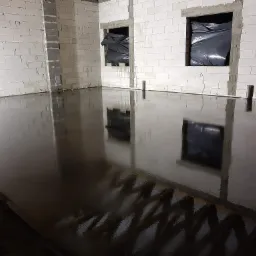 Świeżo wylana, błyszcząca posadzka w surowym wnętrzu z białych bloczków, okna zabezpieczone czarną folią