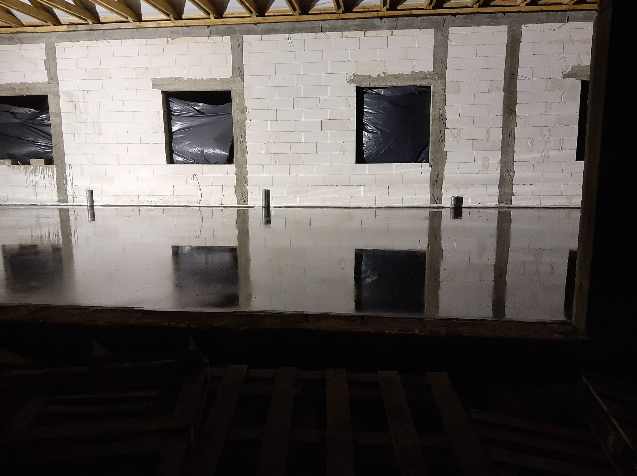 Świeżo wylana, błyszcząca posadzka w surowym wnętrzu z bloczków, okna zabezpieczone czarną folią, widoczne drewniane elementy konstrukcyjne stropu.