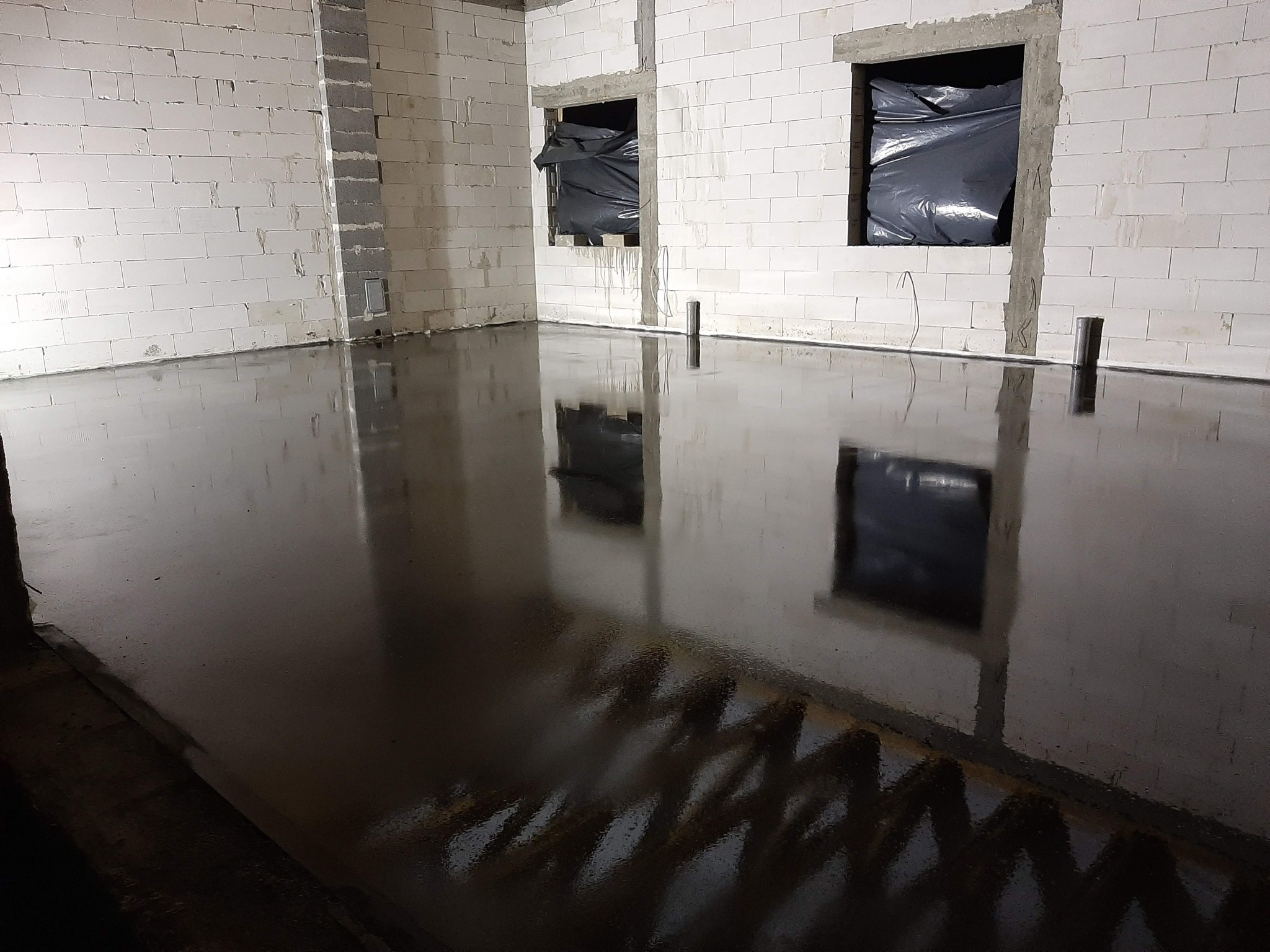 Świeżo wylana, błyszcząca posadzka w surowym wnętrzu z białych bloczków, okna zabezpieczone czarną folią