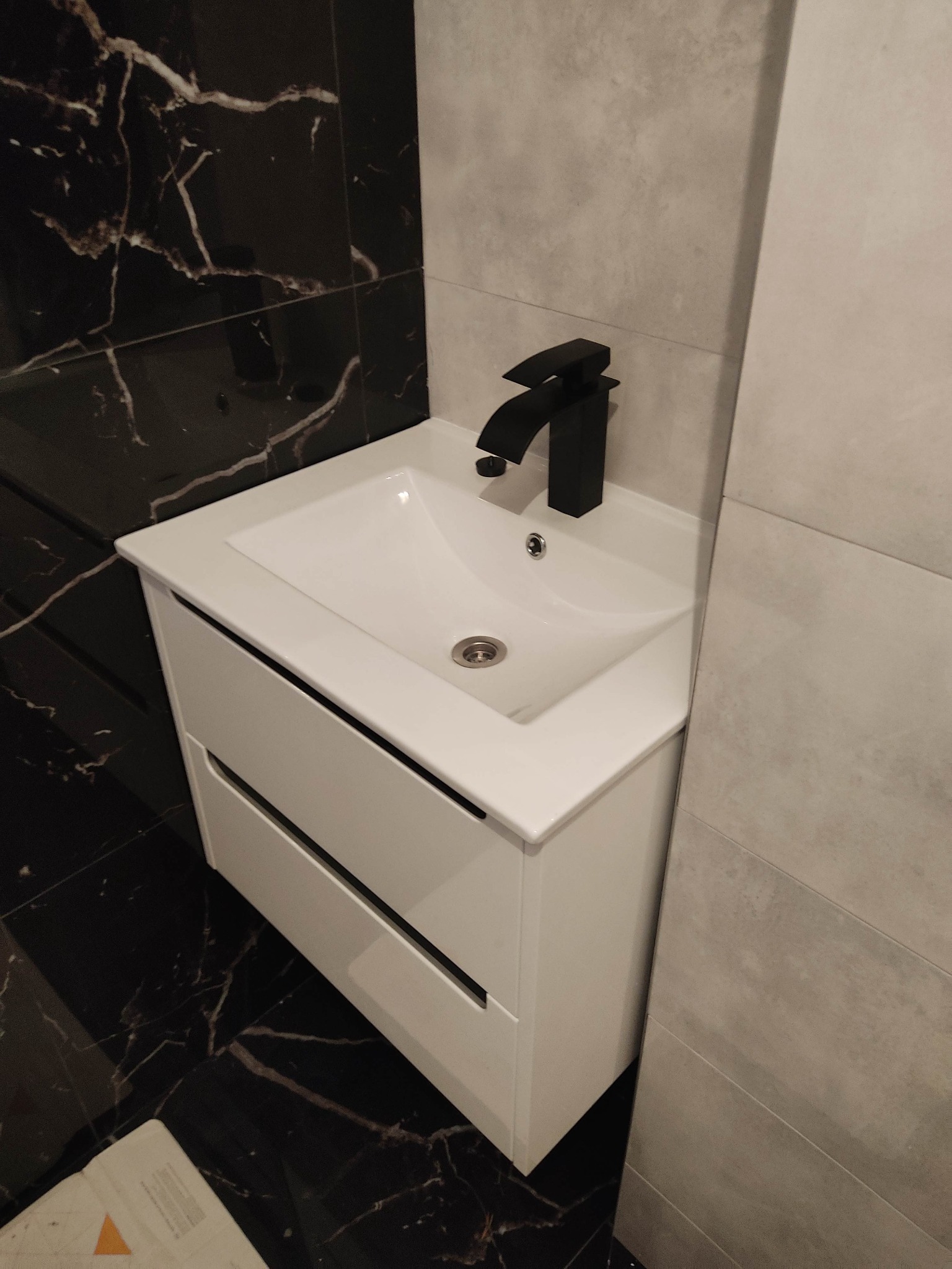 Nowoczesna łazienka z białą szafką pod umywalkę z szufladami i czarną armaturą na tle czarnej ściany imitującej marmur i jasnoszarej ściany z płytek.