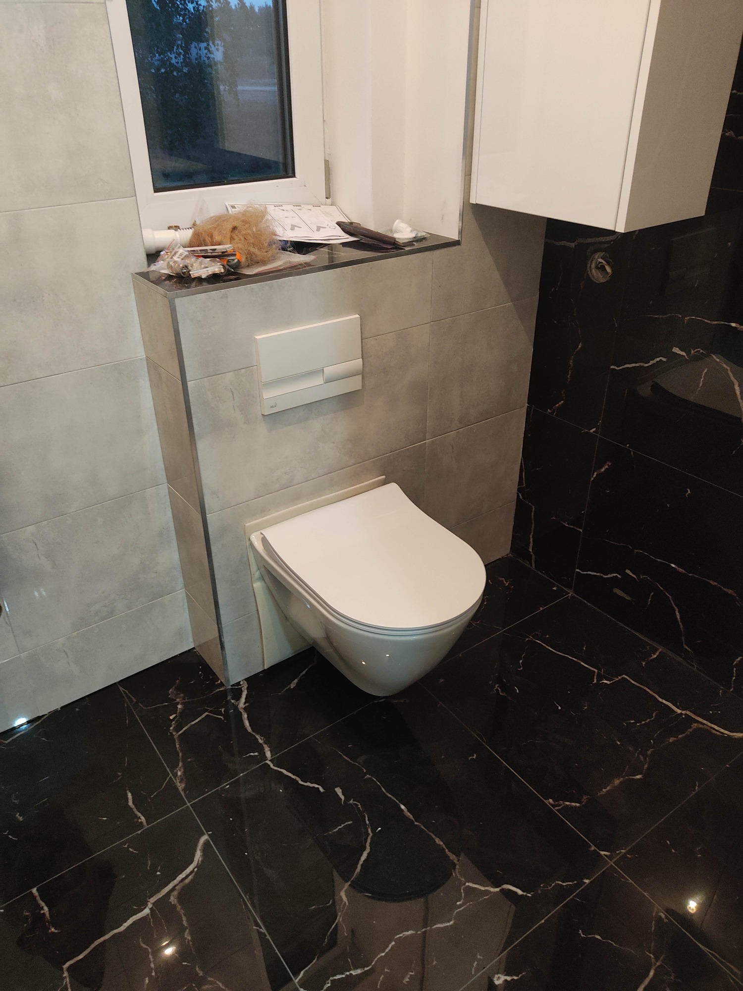 Podwieszana toaleta z szarą zabudową i czarną marmurową podłogą, widoczne narzędzia i instrukcje na półce pod oknem, biała szafka wisząca w rogu