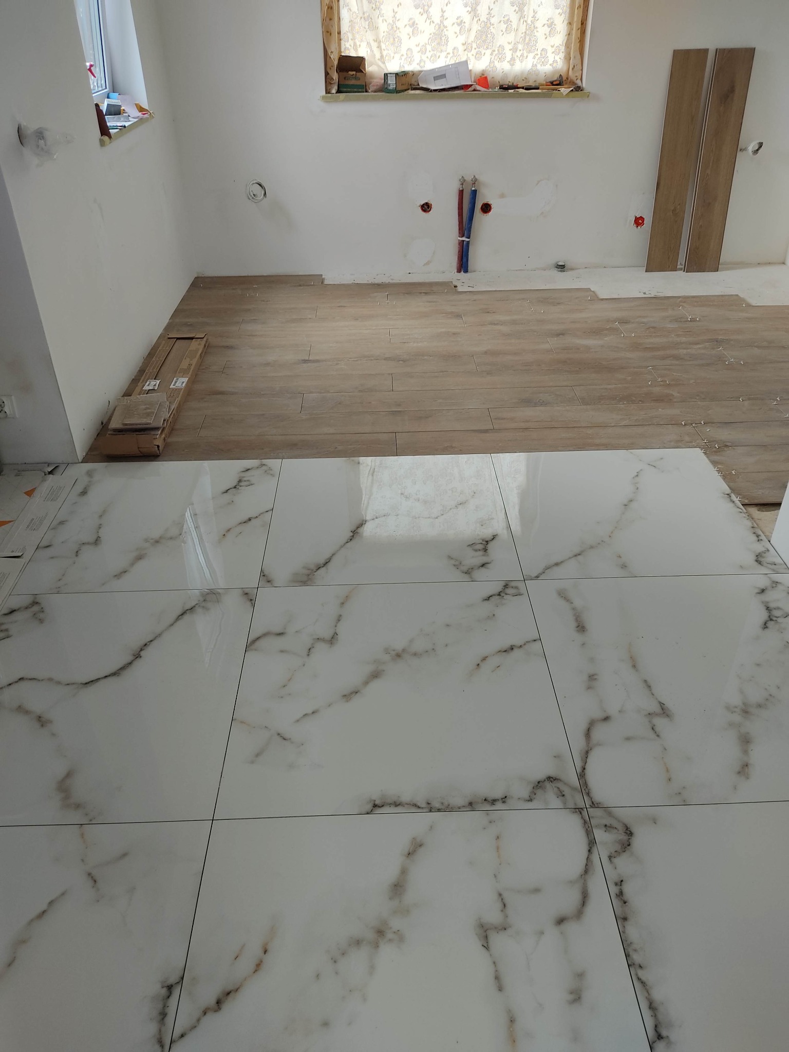 Układanie białych płytek podłogowych z marmurkowym wzorem w pomieszczeniu z częściowo ułożoną drewnopodobną podłogą i przygotowanymi instalacjami hydraulicznymi.