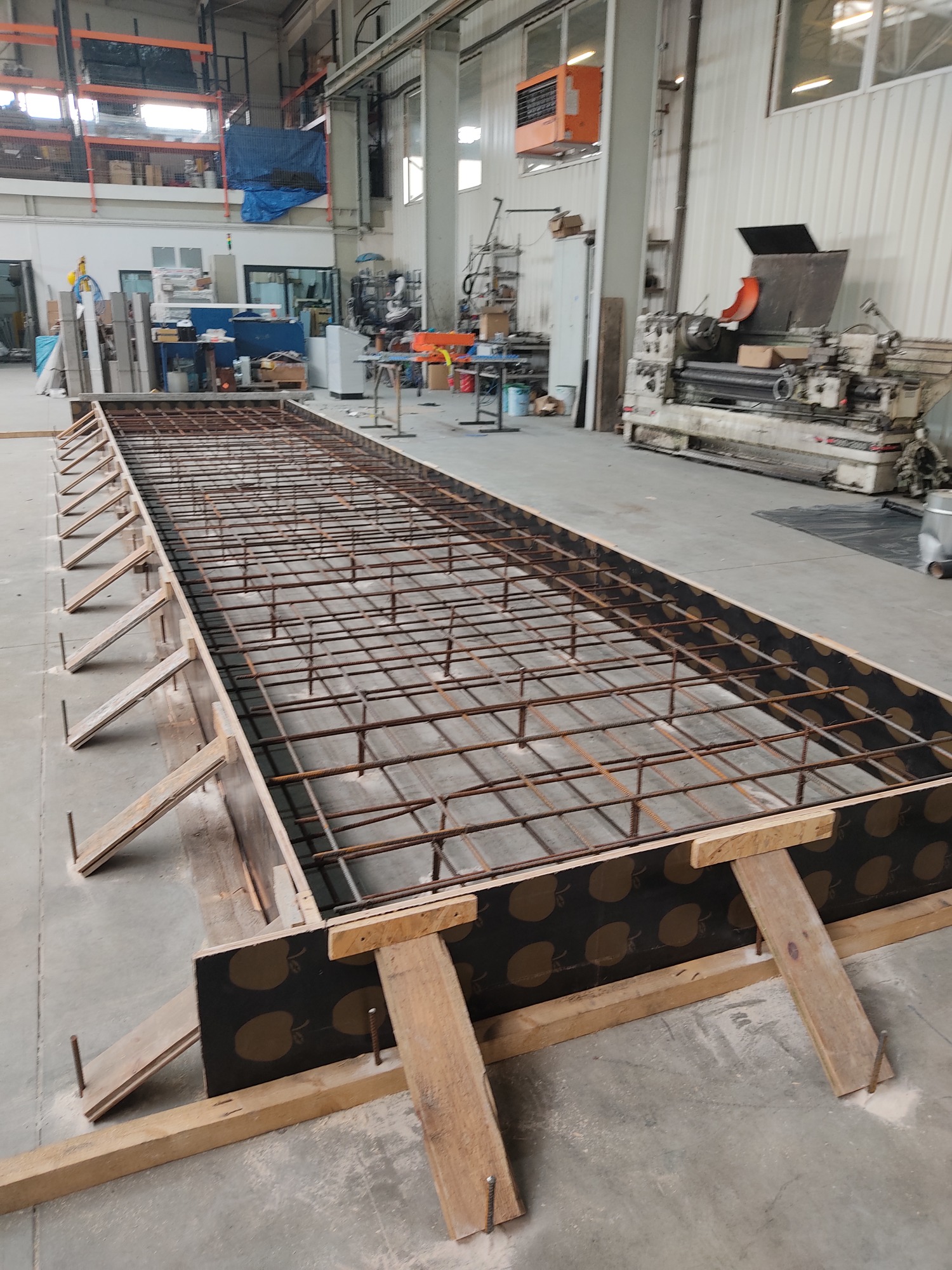 Przygotowanie szalunku z drewnianymi podporami i zbrojeniem z siatki stalowej w hali produkcyjnej, widoczna maszyna tokarska w tle.