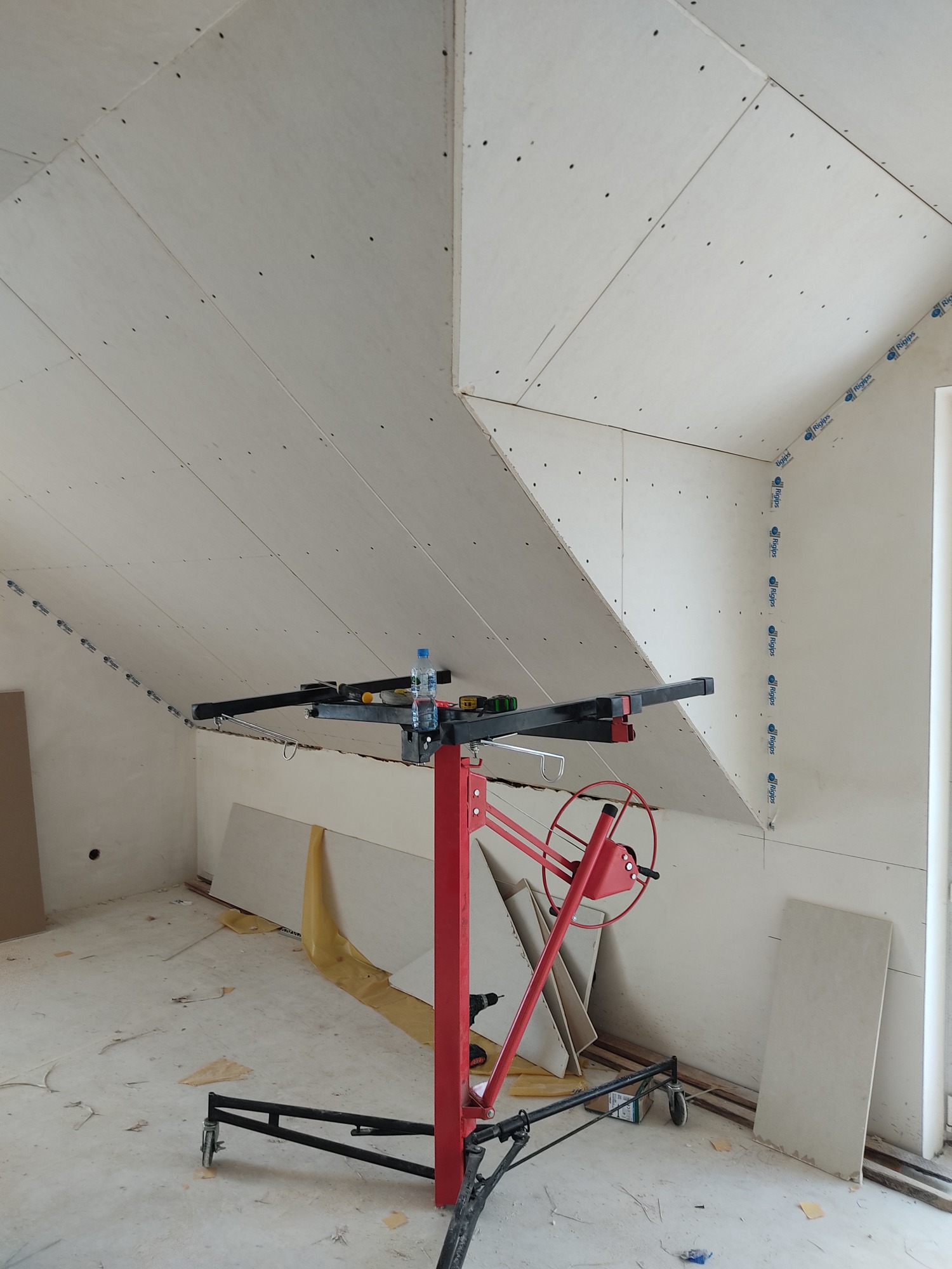 Montaż płyt gipsowo-kartonowych na poddaszu, widoczny podnośnik do płyt, częściowo zabudowane skosy, taśma Rigips na łączeniach ścian, surowy stan wykończenia.