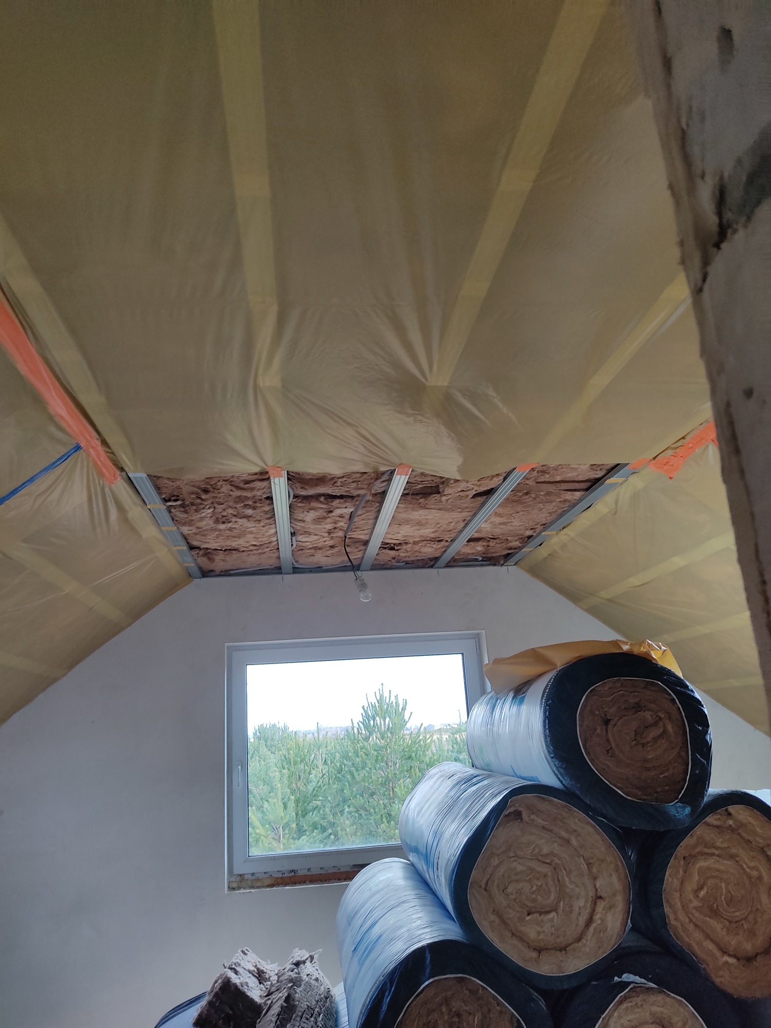 Poddasze w trakcie izolacji: widoczne rolki wełny mineralnej w folii, częściowo ocieplony sufit z widoczną konstrukcją metalową i wystającą żarówką, okno z widokiem na las.